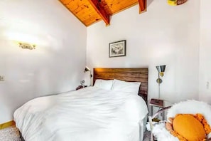A bedroom with a wooden ceiling, a bed with white bedding, a nightstand, a lamp, and a framed picture on the wall.