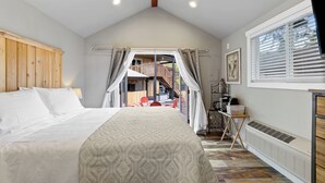 A bedroom with a large bed, a wooden headboard, a window with blinds, a radiator, and a view of an outdoor area with chairs and a table. - Cozy Bungalow W/ Lake Access - Near Mt. Rainier (Eatonville)
