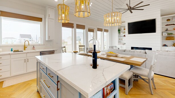 Une cuisine moderne avec des meubles blancs, un îlot en marbre, des luminaires suspendus et un grand téléviseur fixé au mur.