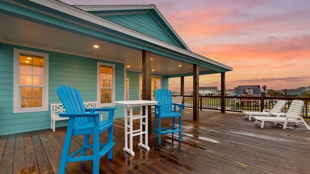 A covered deck with wooden flooring, blue Adirondack chairs, a white table, and a view of the sunset. - Retreat | Gulf Views | Large Deck | Group Ready (Freeport)