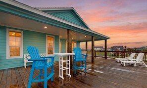 A covered deck with wooden flooring, blue Adirondack chairs, a white table, and a view of the sunset. - Retreat | Gulf Views | Large Deck | Group Ready (Freeport)