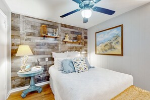Une chambre avec un mur en bois, un ventilateur de plafond, un lit avec une literie blanche, une petite table ronde avec une lampe et un tableau représentant une scène de plage.
