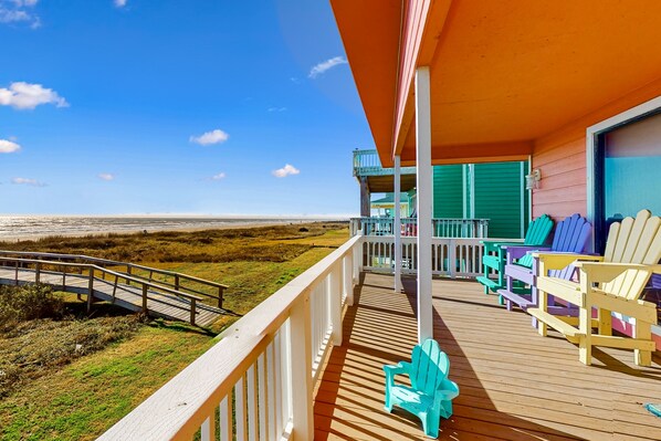 Une terrasse en bois offrant une vue sur la plage, un espace abrité avec des fauteuils Adirondack aux couleurs vives, et un escalier en bois menant à une allée couverte.