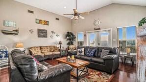 A spacious living room with a ceiling fan, a wooden coffee table, and a large window with blinds. - Retreat | Steps to Beach | Dog Haven | Sleeps 12 (Crystal Beach)