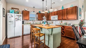 A kitchen with wooden cabinets, a central island, bar stools, a microwave, and a refrigerator. - Retreat | Steps to Beach | Dog Haven | Sleeps 12 (Crystal Beach)
