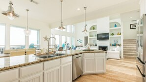 A spacious kitchen with a large island, white cabinetry, and granite countertops. There is a dining area with a white table and chairs, and a living room with a fireplace and a television.
