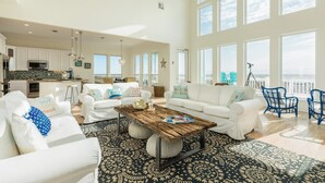 A spacious living area with a large coffee table, white sofas with decorative pillows, and a view of the ocean through large windows.