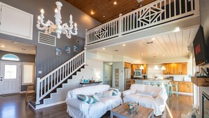Un vaste espace de vie comprenant un escalier, des canapés blancs, une table basse en bois et un lustre.
