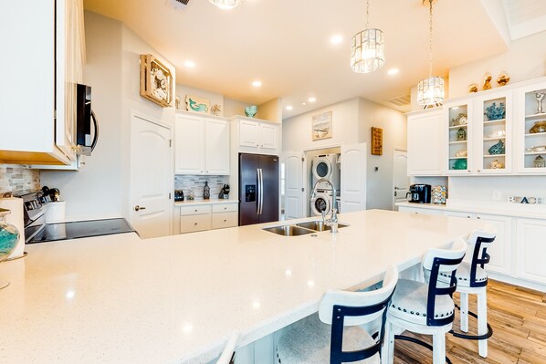 A modern kitchen with white cabinetry, a large island with bar stools, stainless steel appliances, and decorative items on shelves. - Amazing | Gulf Views | Large Deck | Family Fun (Freeport)