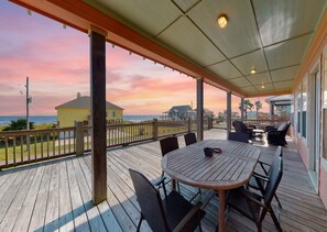 A covered outdoor deck with a table and chairs, overlooking a beach with houses in the background. - Ultimate | Ocean Views | Dog Haven (Crystal Beach)