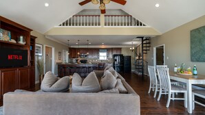 A spacious living area with a large gray sofa, a dining table with chairs, a kitchen in the background, and a staircase leading to an upper level.