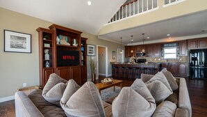 A spacious living room with a large brown sofa, a wooden entertainment center with a Netflix sign, and a kitchen in the background.