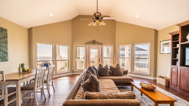 A spacious living room with a large sectional sofa, a dining table with chairs, and a wooden coffee table. There is a ceiling fan, large windows offering a view of the outdoors, and a television on the right side.