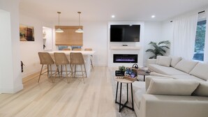 A modern living room with a sofa, a TV, and a fireplace. There is a dining area with a table and chairs. - Retreat by the Vineyard |Panoramic Sauna |Hot Tub |EV Charger (Niagara-on-the-Lake)