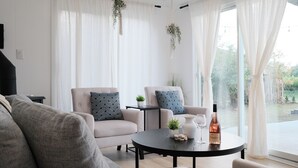 A modern living room with a grey sofa, two armchairs, a small table with a wine bottle and glass, and a view of greenery outside. - Retreat by the Vineyard |Panoramic Sauna |Hot Tub |EV Charger (Niagara-on-the-Lake)