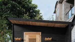 A modern wooden sauna structure with a door and bench, set against a dark exterior wall, with a basket of towels on the ground. - Retreat by the Vineyard |Panoramic Sauna |Hot Tub |EV Charger (Niagara-on-the-Lake)