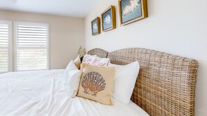 Un dormitorio con una cama grande, ropa de cama blanca, un cabecero de mimbre y tres cuadros en la pared.