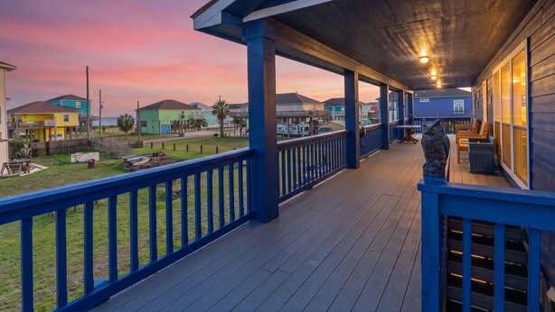 Una terraza de madera con baranda, con vista a un barrio residencial con casas coloridas y un cielo al atardecer.