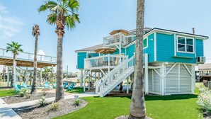A turquoise two-story building with a white balcony and stairs, surrounded by palm trees and outdoor seating. - Dream | Canal Front | Firepit | Kayking & Fishing (Surfside Beach)