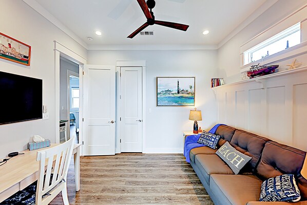 A living room with a brown leather sofa, a flat-screen TV, a ceiling fan, and a painting of a coastal scene.
