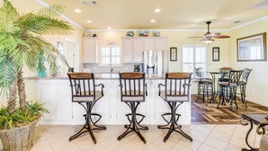 A kitchen with white cabinets, a central island with bar stools, a dining area with a table and chairs, and a ceiling fan. - Perfect | Steps to Beach | Firepit | Sleeps 10 (Crystal Beach)