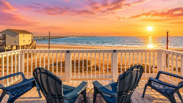 A beach view with a wooden deck, chairs, and a pier in the background.