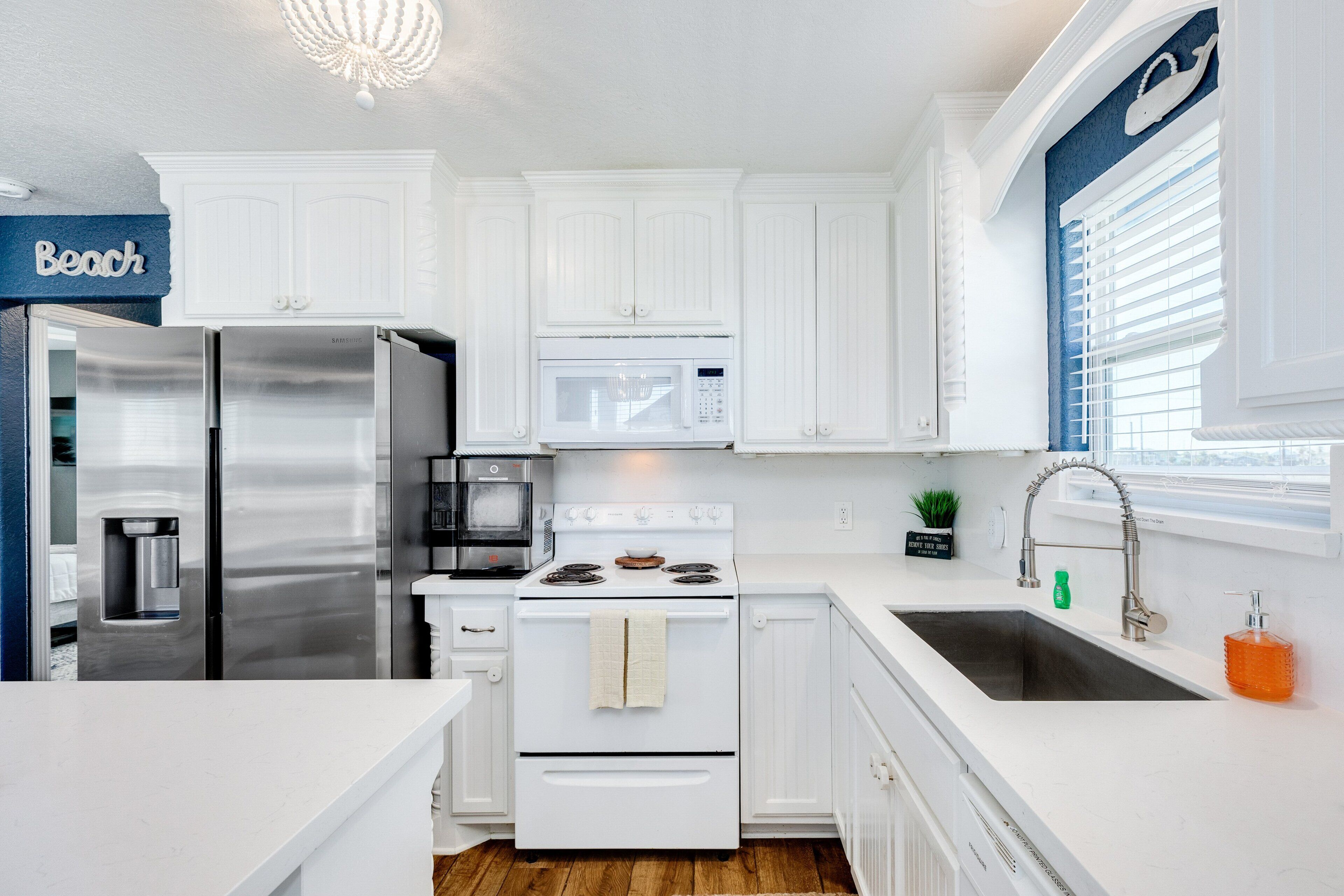 Una cocina moderna con armarios blancos, electrodomésticos de acero inoxidable y vistas a la playa a través del ventanal.