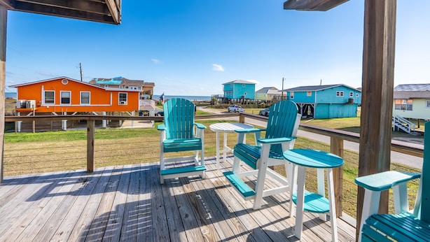 A wooden deck with turquoise chairs and tables, overlooking a beach with colorful houses. - Retreat | Gulf Views | Large Deck | Family Fun (Surfside Beach)
