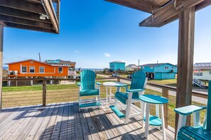 Una terraza de madera con sillas y mesas turquesas, con vista a una playa y casas coloridas.