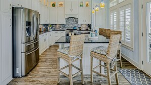 A spacious kitchen with white cabinetry, a stainless steel refrigerator, a large island with a sink, and two wicker chairs.