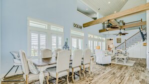 A spacious dining area with a wooden table and chairs, large windows, and a staircase leading to an upper level.