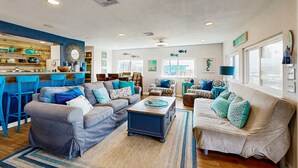 A spacious living room with a grey sofa, a beige sofa, and a coffee table. There are blue chairs and a kitchen area with a bar counter. - Haven | Ocean View | Large Deck | Pet Friendly (Surfside Beach)