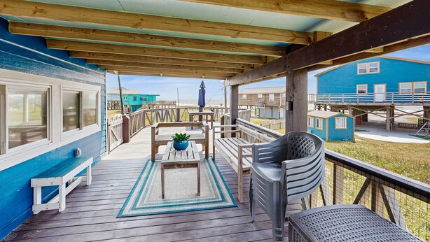 A covered outdoor deck with wooden beams, a blue building, and a small blue structure in the background. - Haven | Ocean View | Large Deck | Pet Friendly (Surfside Beach)