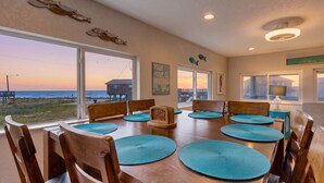 A dining area with a view of the beach, wooden chairs, blue placemats, and a sign that reads 'YOUR HOME IS WAITING.' - Haven | Ocean View | Large Deck | Pet Friendly (Surfside Beach)