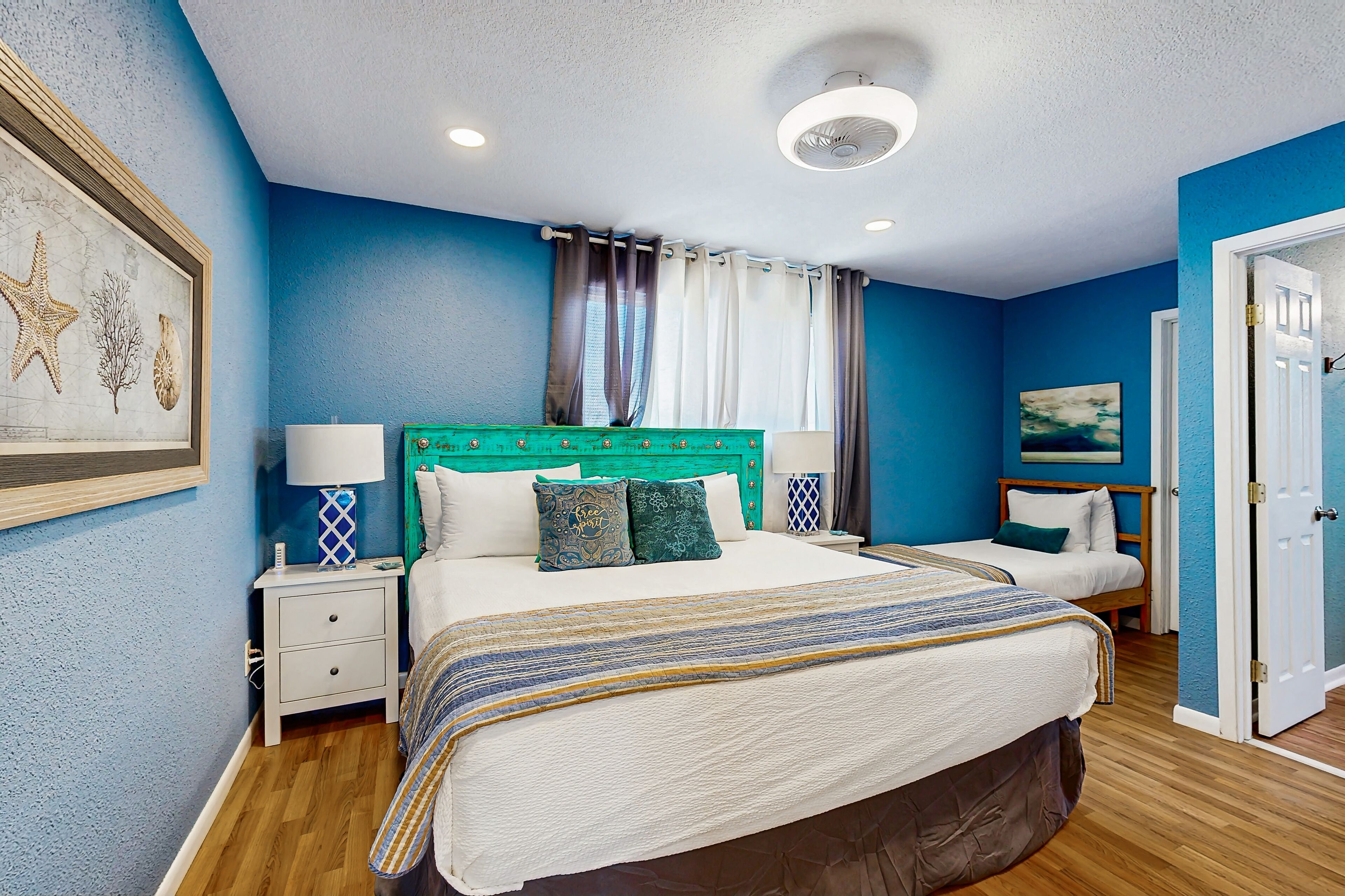 A bedroom with two beds, a teal headboard, white bedding, a framed seashore artwork, and a window with curtains.