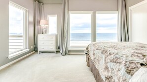 A bedroom with a large bed, a dresser, a lamp, and a view of the ocean through large windows. - Beachfront 4br, Hot Tub, Multiple Deck, Sleeps 16 (Gilchrist)