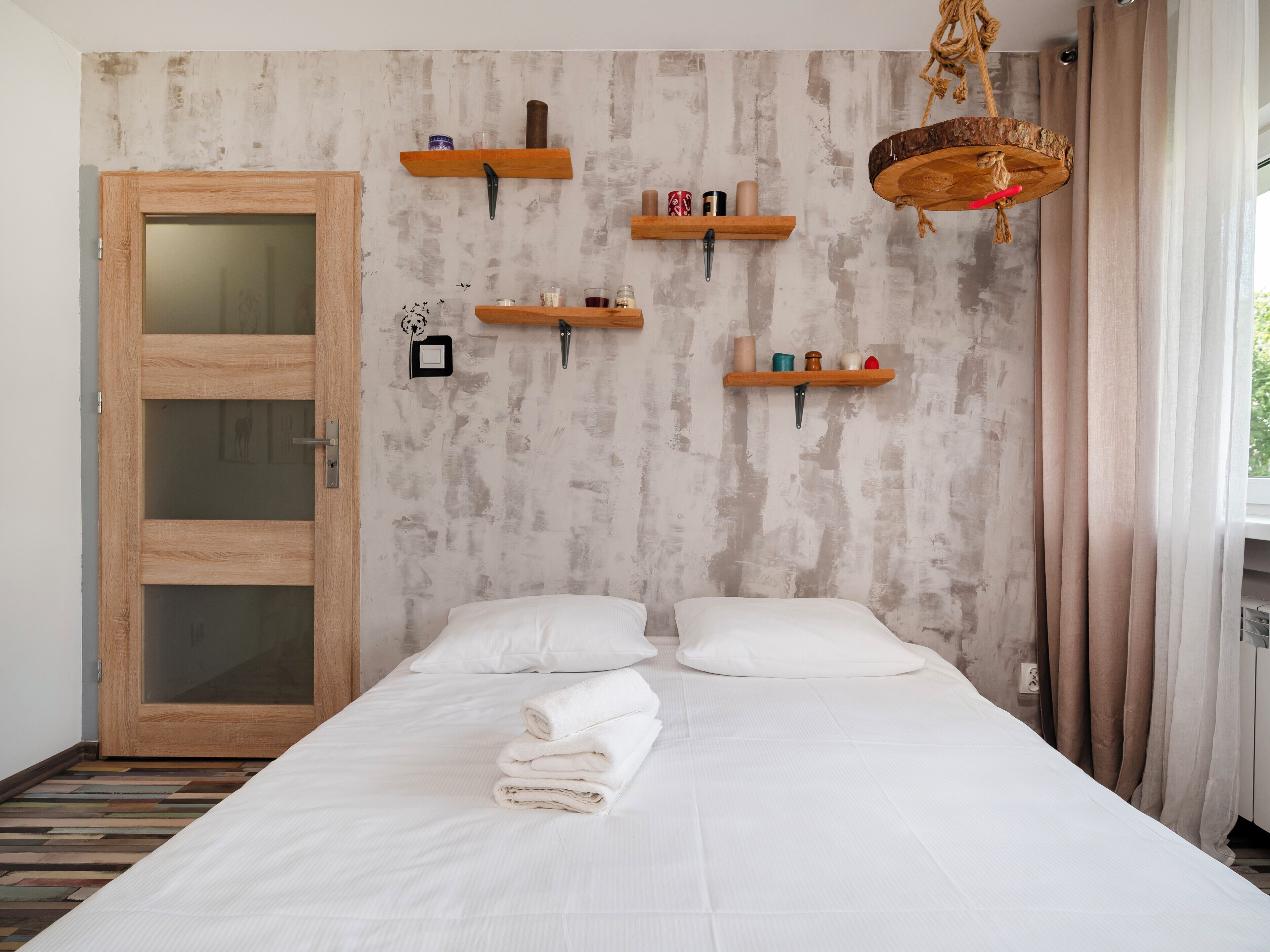 A bedroom with a wooden door, a bed with white linens, and multiple wooden shelves on the wall.