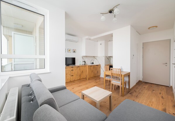 A modern living room with a gray sofa, a wooden dining table, and a kitchen area with wooden cabinets and a TV.