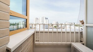 Un balcón con vistas a edificios de la ciudad, barandal blanco y ventana con marco de madera.