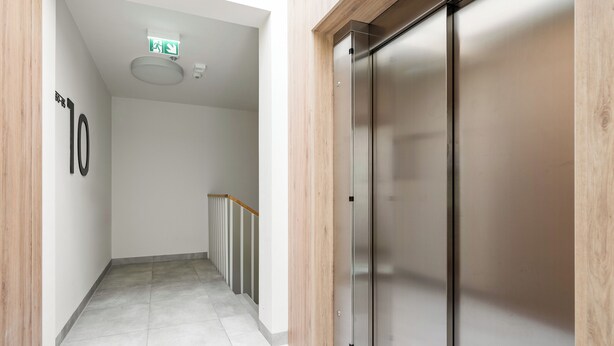 Un pasillo moderno con un ascensor de acero inoxidable, piso de madera y pared blanca con un letrero de número.