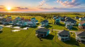 A residential area with houses featuring multiple stories and attached porches, set against a backdrop of a sunset over a coastal landscape. - Prime | Gulf Views | Large Deck | Pet Paradise (Crystal Beach)