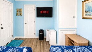 A room with two beds, a television displaying 'NETFLIX', a vacuum cleaner, a washing machine, and a small wooden table.