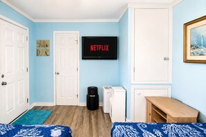 A room with two beds, a television displaying 'NETFLIX', a vacuum cleaner, a washing machine, and a small wooden table.