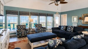A living room with a sofa set, a coffee table, and patterned cushions. There is a ceiling fan, a door with a sign, and a view through large windows.