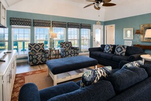 A living room with a sofa set, a coffee table, and patterned cushions. There is a ceiling fan, a door with a sign, and a view through large windows.