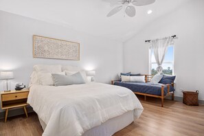 Une chambre avec un grand lit, un lit plus petit, une table de chevet, une œuvre d’art murale, une fenêtre avec des rideaux et un ventilateur de plafond.