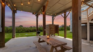 Eine Terrass mit Holz-Picknicktischen und Lichterketten, mit Blick auf eine Rasenfläche und ein Haus.