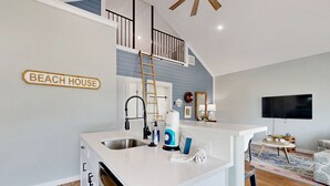 Eine moderne Küche mit weißer Arbeitsplatte, Spüle aus Edelstahl und einer Holzleiter. Ein Schild an der Wand zeigt den Schriftzug "BEACH HOUSE".