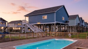 Ein Poolbereich mit Holzterrasse, ein blaues Haus mit Balkon und ein klarer Himmel.