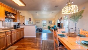 Une cuisine spacieuse avec des meubles en bois, un coin repas avec une table dressée pour quatre personnes, et un salon à l’arrière-plan.